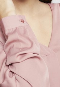 Blusa di raso rosa caratterizzata da maniche lunghe e gonfie con un bottone decorativo sul polsino e un leggero scollo a V. Texture liscia.