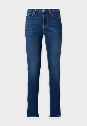 Donna con capelli lunghi e scuri che indossa una canotta nera, jeans skinny blu e ballerine nere, in piedi davanti a uno sfondo semplice.