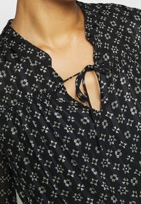 Blouse noire à motifs avec des dessins géométriques d'étoiles blanches, tissu froncé et un lien devant au niveau du col.