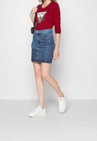 Röd långärmad skjorta med en logografik, i kombination med en blå denimpennkjol. Modellen bär vita sneakers och står på en neutral bakgrund.