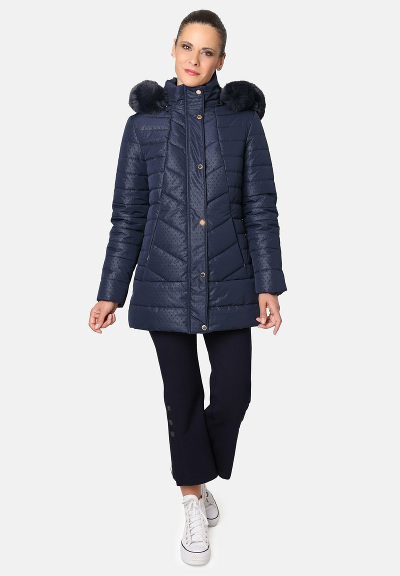 Abrigo acolchado azul marino con patrón en forma de chevron, capucha con borde de piel, cierre de cremallera y botones, combinado con pantalones oscuros y zapatillas blancas.