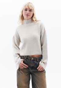 Hellbeige, gerippter Rollkragenpullover mit Ballonärmeln und verkürztem Design. Kombiniert mit hochtaillierten, ausgewaschenen Jeans.