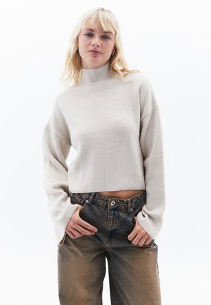 Hellbeige, gerippter Rollkragenpullover mit Ballonärmeln und verkürztem Design. Kombiniert mit hochtaillierten, ausgewaschenen Jeans.