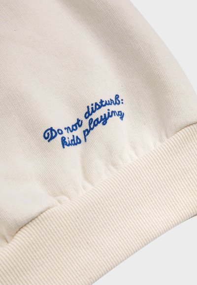 Felpa cremosa con polsini a coste. Il testo ricamato in blu recita: "Non disturbare: bambini che giocano." Tessuto dal tocco morbido.
