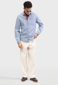 Chemise à rayures bleues et blanches avec une poche avant, assortie à un pantalon large couleur crème et des chaussures marron foncé. Accent en bandana rouge.
