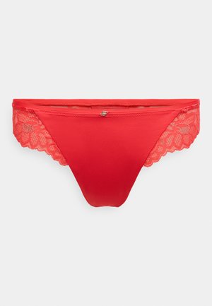 Sarkanas tangas ar mīkstu audumu, mežģīņu sānu paneļiem, viļņotām malām un nelielu dekoratīvu akcentu priekšējā centrā.