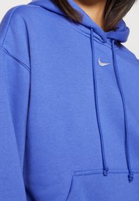 Blå sweatshirt med en stor kanguruficka och huva med dragsko. Har en liten metallisk Nike-logga på bröstet. Mjukt, texturerat tyg.