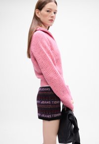 Roze pleteni pulover s teksturiranim uzorkom, uparen s tamnim pletenim šortsevima s printom logotipa. Model drži crnu torbu.