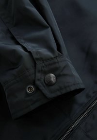 Zwarte waterdichte jasmanchet met een knoop- en ringdetail, een gladde textuur en een zichtbare ritssluiting langs de rand.