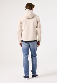 Lichtbeige hoodie met een gestructureerd ontwerp, voorzien van een capuchon en zwarte voering. Draagt bij blauwe denim jeans en casual marineblauwe sneakers.