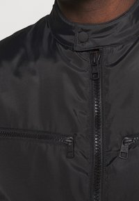 Giacca leggera nera con colletto alto, dotata di una cerniera centrale e due tasche con cerniera, realizzata in materiale sintetico liscio.