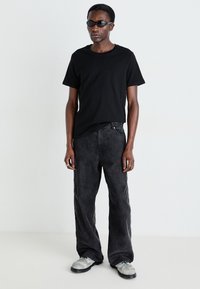 T-shirt en coton noir associé à un pantalon gris foncé ample. Les accessoires comprennent des bagues en argent et des lunettes de soleil sombres. Les chaussures sont des chaussures grises texturées.