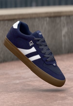 Navy suede sneaker with white stripes, grey laces, and a gum sole. Features padded collar and logo on tongue. Casual and durable design.