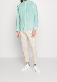 Camicia in lino turchese con colletto alla mandarina, chiusura con bottoni, abbinata a pantaloni tapered beige chiaro e sneakers bianche.