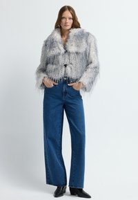 Hüftkurz geschnittener Mantel mit einer flauschigen grau-weißen Textur, ausgestattet mit einem großen Kragen und fransigen Kanten. Kombiniert mit hoch taillierten blauen Jeans und schwarzen Stiefeln.