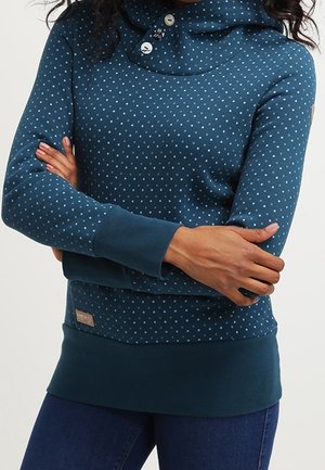 Femme portant un sweat à capuche à manches longues couleur sarcelle avec des pois bleu clair, bras croisés et jean bleu foncé sur un fond uni.