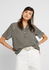 Chemise à manches courtes et à boutons de couleur vert foncé avec un motif géométrique. Associée à un pantalon blanc et une ceinture de couleur claire.