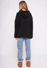 Czarna dzianinowa bluza z kapturem o fakturowanym wykończeniu, duży kaptur i żebrowany dół, zestawiona z jasnoniebieskimi dżinsami i brązowymi butami ocieplanymi futerkiem.