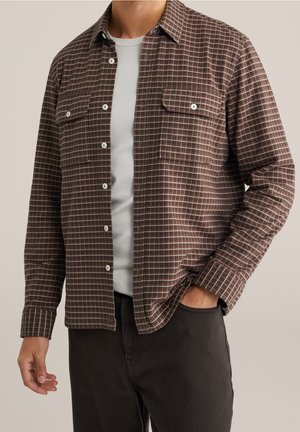 Muž v hnědé kostkované košili na knoflíky přes bílé tričko a černé kalhoty, jedna ruka v kapse, neutrální pozadí.