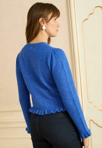 Nő oldalra fordulva, kék hosszú ujjú pulóvert visel fodros alsó résszel és fekete nadrágot, krémszínű, díszléccel ellátott fal mellett áll.