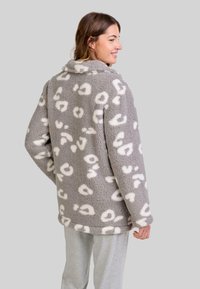 Mujer vistiendo una chaqueta de forro polar gris con un patrón de leopardo blanco, combinada con pantalones gris claro, de perfil y sonriendo.