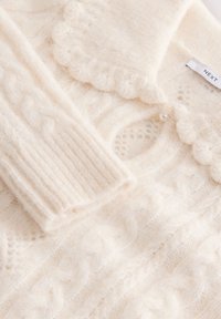 Kremno obarvan pulover iz pletenine z vzorcem kablov, nazobčenim ovratnikom, rebrastimi manšetami in zapiranjem na gumbe, okrašenim z majhno biserno kroglico.