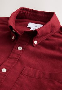 Chemise bordeaux à boutons en tissu doux, avec col, poche et boutons blancs. Étiquette indiquant "NEXT". Design simple et structuré.