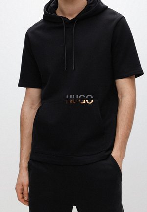 Mies pukeutuneena mustaan lyhythihaiseen hupulliseen vetoketjulliseen collegepaitaan, jossa metallinen "HUGO"-logo, ja mustiin housuihin, kädet taskuissa, valkoinen tausta.