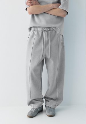 Persona che indossa pantaloni della tuta larghi grigio chiaro, t-shirt abbinata e scarpe da ginnastica grigie, in piedi con le braccia incrociate contro un muro bianco.