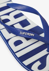 Mėlynos gumos šlepetės su tekstūruotu pado ir dirželio paviršiumi, su "SUPERDRY" ženklu ir ryškiai balta užrašų juosta ant pado.