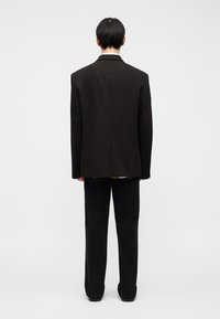 Άτομο με κοντά μαύρα μαλλιά στέκεται γυρισμένο προς τα πίσω, φοράει μια υπερμεγέθη μαύρη σακάκι, μαύρο παντελόνι και μαύρα παπούτσια μπροστά σε α πλό φόντο.