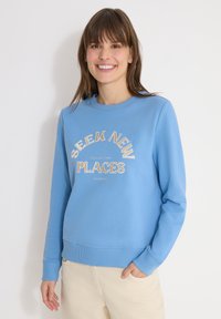 Femme souriante aux cheveux bruns portant un sweat-shirt bleu clair avec le texte "SEEK NEW PLACES COLLECTING STORIES" et un pantalon beige, main dans la poche.