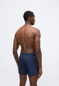 Tmavě modré koupací šortky s elastickým pasem, vybavené zadní kapsou a červeným logem. Lehká tkanina s hladkou texturou.
