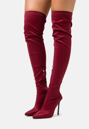 Botas de tacón stiletto de color burdeos hasta la rodilla, con un tejido elástico y unos detalles de cremallera lateral, que presentan una superficie suave y mate.
