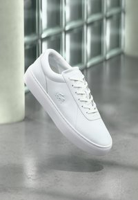 Λευκό χαμηλό κλασικό παπούτσι με δέσιμο κορδονιών και ανάγλυφο λογότυπο κροκόδειλου, τοποθετημένο πάνω σε γκρίζο γυαλιστερό πάτωμα με φόντο τοίχο από γυάλινους κύβους.