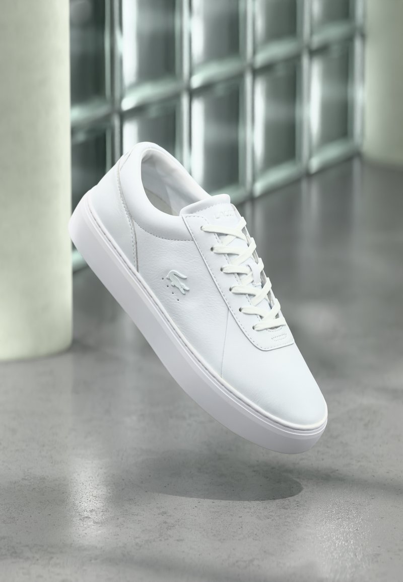 Λευκό χαμηλό κλασικό παπούτσι με δέσιμο κορδονιών και ανάγλυφο λογότυπο κροκόδειλου, τοποθετημένο πάνω σε γκρίζο γυαλιστερό πάτωμα με φόντο τοίχο από γυάλινους κύβους.