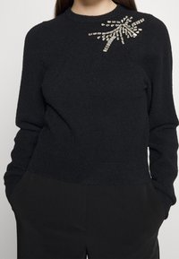 Czarny sweter dzianinowy z długimi rękawami, z ozdobnymi srebrnymi elementami w kwiatowy wzór na górnej lewej stronie.
