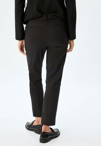 Pantalons noirs ajustés avec une texture lisse, dotés d'une taille mi-haute et de deux poches arrière, associés à des chaussures noires brillantes à enfiler.