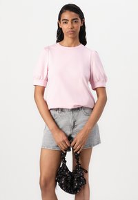 Rosa, kortärmad topp med puffärmar, matchad med grå denimshorts. Modellen håller en svart blommig scrunchie-väska.