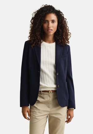 Femme aux cheveux bouclés portant un blazer bleu marine, un pull en maille torsadée blanc, un pantalon beige et une ceinture marron, debout devant un fond uni.