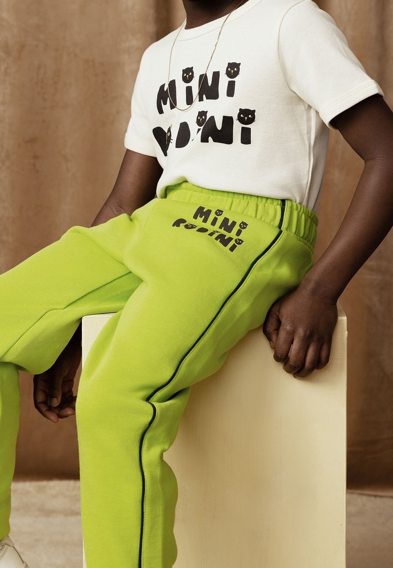 Enfant assis sur une boîte en bois clair, portant un t-shirt blanc "Mini Rodini" et un pantalon de survêtement vert vif avec des lettres et des accents noirs.