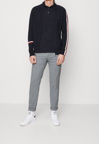 Giacca di felpa zip blu navy con strisce bianche e rosse, abbinata a una polo scura e a pantaloni grigi a fantasia; completata con sneakers bianche.