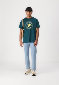 Camiseta de algodón color teal con gráfico circular amarillo "Converse All Star", combinada con jeans azul claro y zapatillas de deporte beige. Bolsa cruzada color crema.