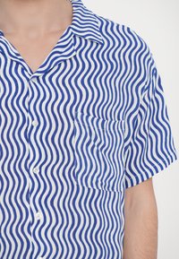 Rövid ujjú, gombos ing kék és fehér hullámmintával, mellzsebbel és lazább gallér kialakítással.