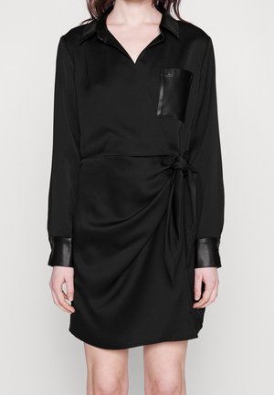 Mujer vestida con un vestido cruzado negro con cuello, bolsillo y puños de cuero, anudado en la cintura, de pie contra un fondo liso.
