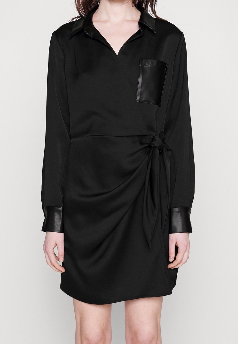 Femme portant une robe portefeuille noire avec un col, une poche et des poignets en cuir, nouée à la taille, debout devant un fond uni.