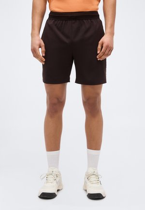 Meesmodell, kes kannab musta sportlühikeste pükse, valgeid sokke ja valgeid tossusid, seisab ühevärvilise heleda taustaga.