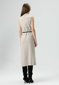 Beige mouwloze jurk met een gestructureerde afwerking, voorzien van een bruine riem in de taille, tot op de knie, en een hoge halslijn.