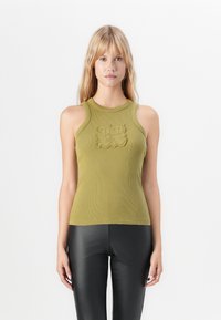 Blond kvinna som bär en ärmlös olivgrön ribbad topp med präglad design, ihop med svarta skinnbyxor, stående mot vit bakgrund.