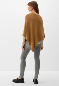 Poncho in maglia marrone con una vestibilità rilassata, caratterizzato da maniche ampie e un orlo asimmetrico, abbinato a jeans aderenti grigi chiari e scarpe nere.